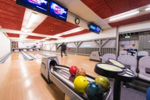Bowlingkuler i forskjellige farger, i bakgrunnen en mann som kaster en bowlingkule på en av tre bowlingbaner.