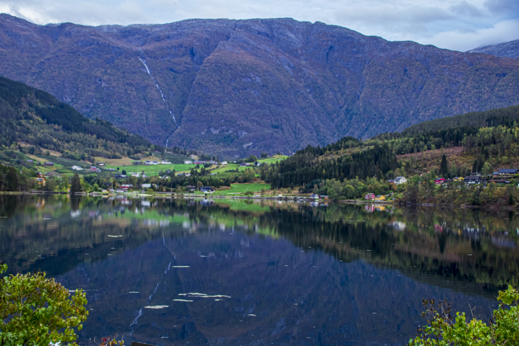 Naturbilde. På andre siden av et vann ser en deler av bygden Ulvik. Grønn mark og fjell i bakgrunnen.