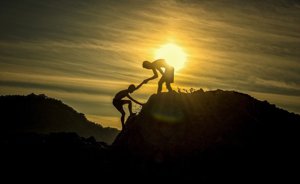 Silhuett av en person som står på toppen av et fjell og strekker armen ut for å hjelpe en person til å komme seg til toppen. I bakgrunnen en sterk sol