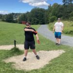 Gutt 18år i svart tøy og solbriller står klar til å kaste en frisbee. Far står i bakgrunnen og observerer.