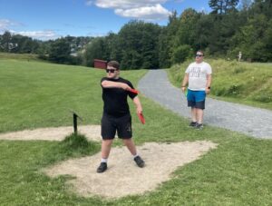 Gutt 18år i svart tøy og solbriller står klar til å kaste en frisbee. Far står i bakgrunnen og observerer.
