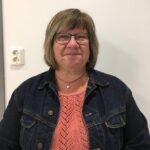 Synnøve Bratlie. Kvinne med brunt hår og briller smiler mot kamera. Har på seg dongerijakke og lys genser