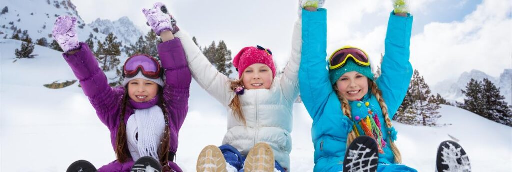 3 jenter sitter i snøen med hendene i været og smiler. De har vinterklær og alpinbriller på hodet.
