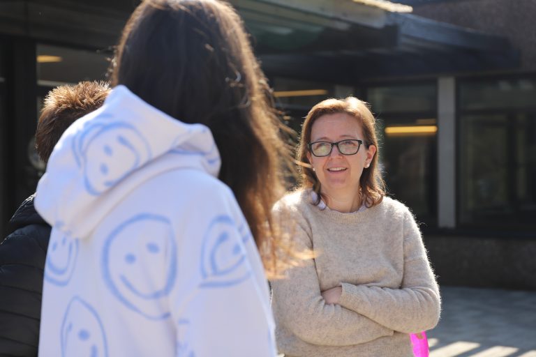 Voksen dame med briller prater med to ungdommer som står med ryggen mot kamera.