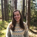 Bilde tatt i skogen. Kvinne med langt brun hår smiler mot kamera. Hun har på seg en strikket grå genser med gult og blått mønster.