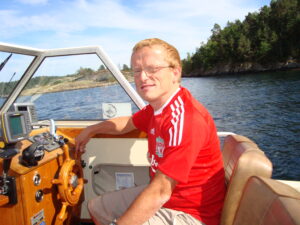 Man i rød t-skjorte ser mot kamera. Han sitter i en båt. I bakgrunnen ser en sjøen og grønne trær