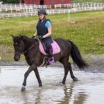 Jente 17år sittende på en svart hest som går i grunt vann. Ved vannkanten går en annen ung jente.
