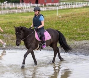 Jente 17år sittende på en svart hest som går i grunt vann. Ved vannkanten går en annen ung jente.