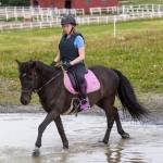 Jente 17år sittende på en svart hest som går i grunt vann. Ved vannkanten går en annen ung jente.
