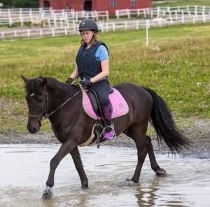 Liv på hesteryggen Jente 17år sittende på en svart hest som går i grunt vann. Ved vannkanten går en annen ung jente.