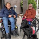 To blide damer i rullestol utenfor Pers Hotell på Gol. Foran svart labrador med rød Servicehund vest.