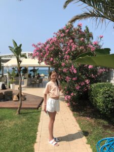 Tenåringsjente i sommerantrekk smilende foran en palme og en busk med rosa blomster.