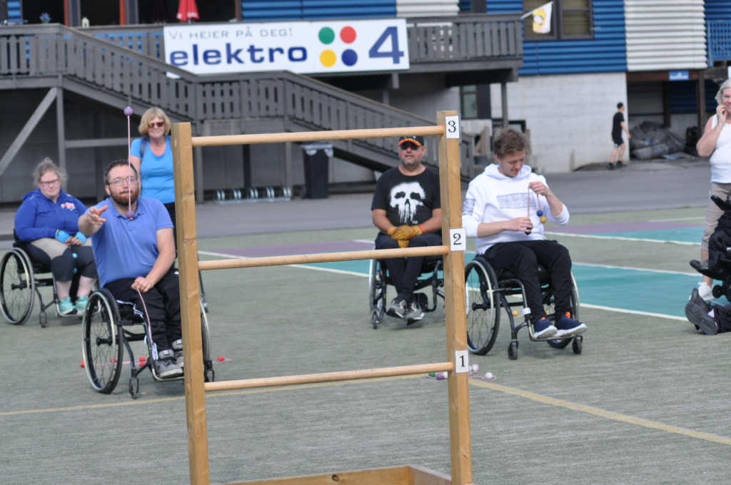 Ung mann som deltar på aktiv sommerferie i Skien Fritidspark. Sitter i rullestol og kaster ball mot stigegolfstativ. Rund sitter og står 5 tilskuere/deltakere.