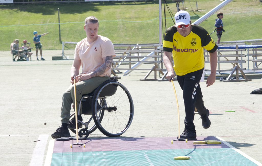 En mannlig rullestolbruker med rosa skjorte og en mann i gul t-skjorte og hvit caps står og ser på et stort Shuffleboard på bakken. Solen skinner.