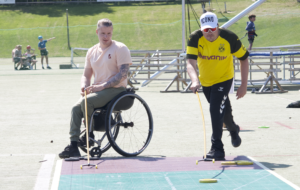 En mannlig rullestolbruker med rosa skjorte og en mann i gul t-skjorte og hvit caps står og ser på et stort Shuffleboard på bakken. Solen skinner.