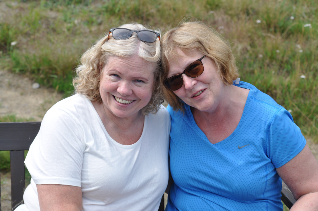 To damer med lyst hår og solbriller smiler mot kamera