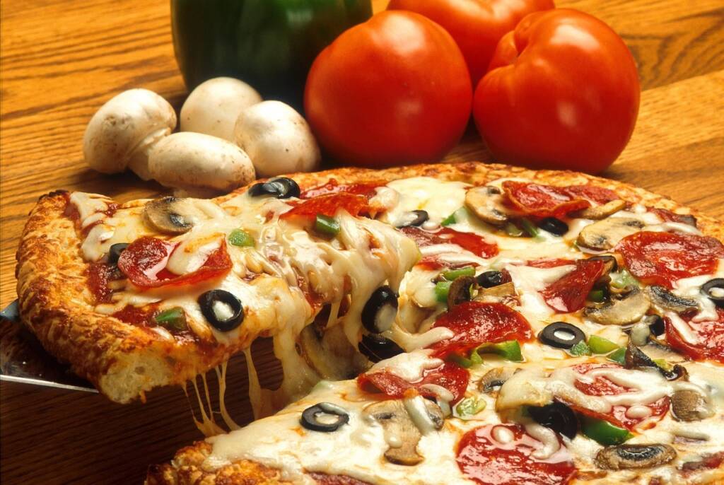 Pizza med pepperoni, paprika og sort oliven. I bakgrunnen er fire champignon og tre tomater.