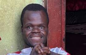 Ung mann i Zambia med hydrocephalus, sitter smilende foran inngangen til huset sitt.