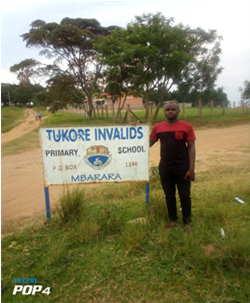 Ung mann som er lærer i Uganda står foran et skilt med skrift "Tukore Invalids - Primary School"