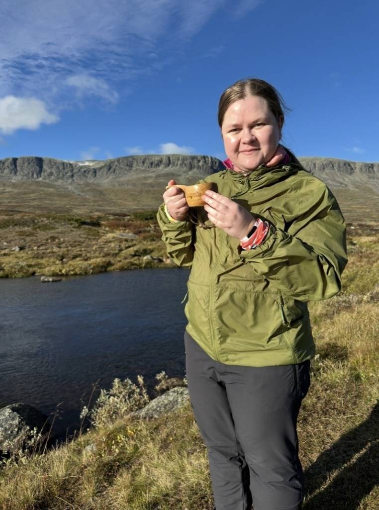 Julie Therese Larsen 4 Bilde av Julie Therese Larsen. En ung dame med blondt hår samlet i hestehale. Står smilende på fjellet i grønn vindjakke og en liten trekopp i hånden.